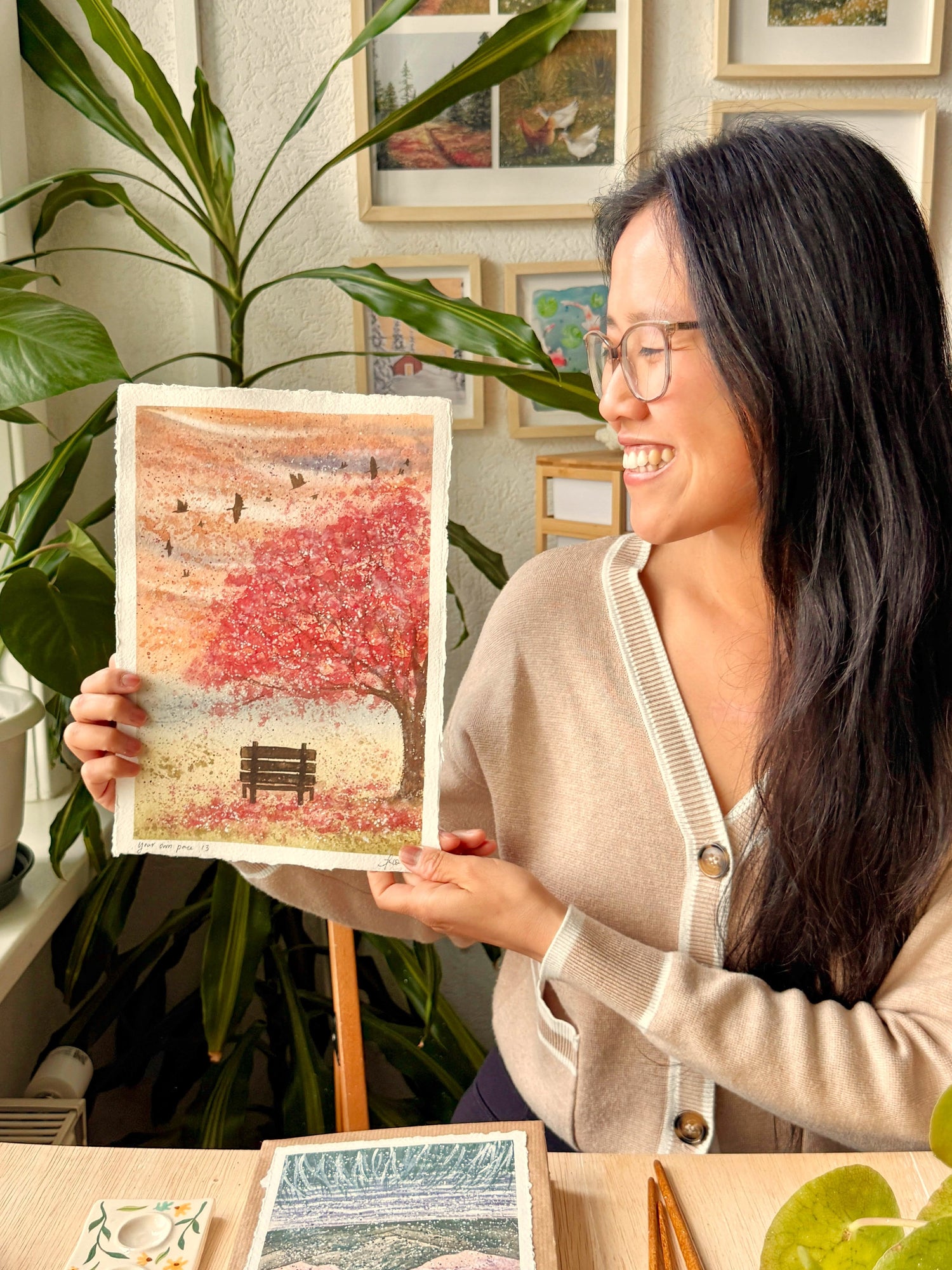 A woman with long dark hair and glasses holds up a colorful art print depicting a red autumn tree and a bench. She is smiling in a decorated room with framed pictures on the wall, potted plants, and other art pieces on a table in front of her.